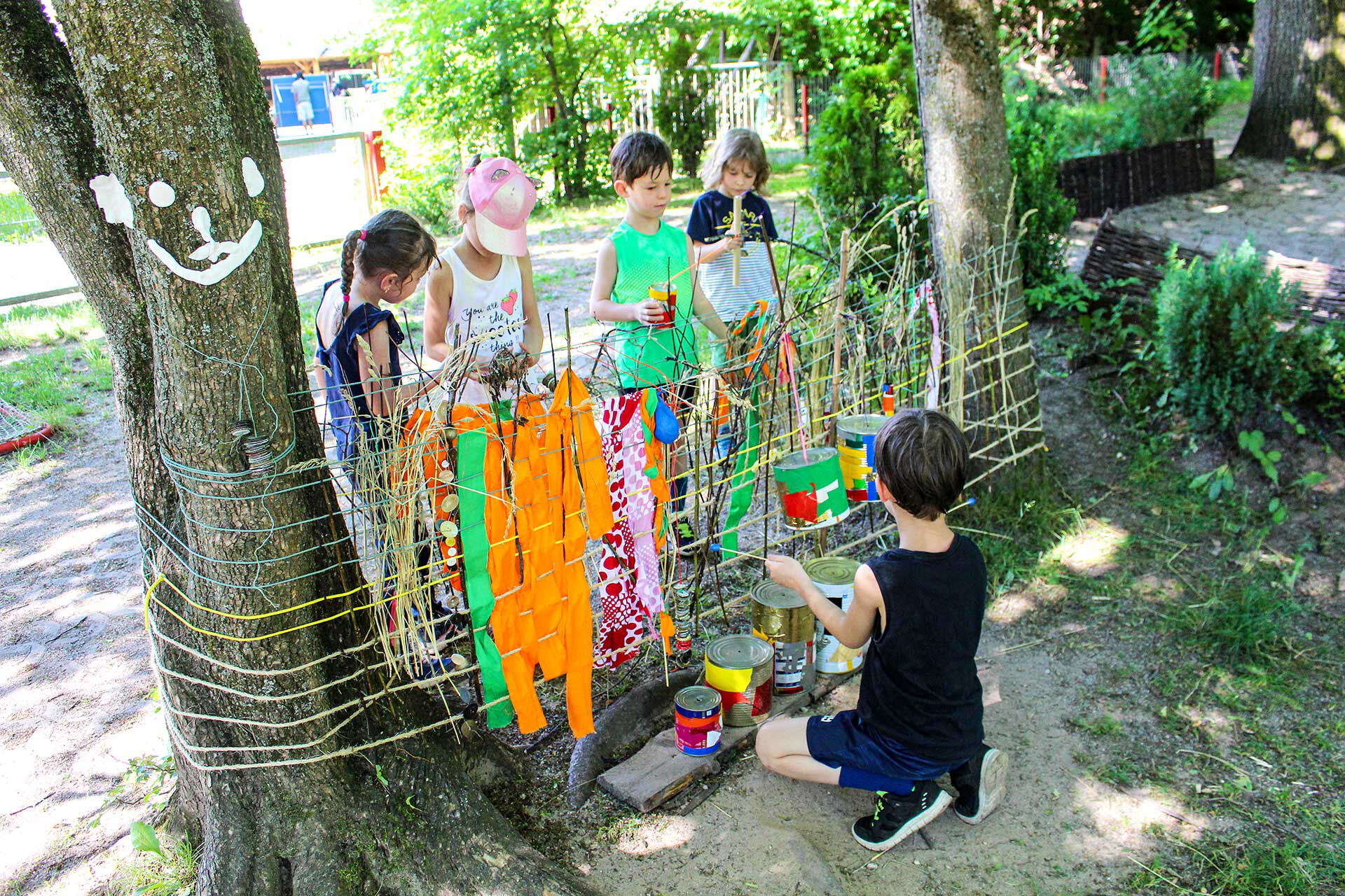 Zwischen zwei Bäumen im Außengelände sind Seile gespannt und Kleidung darüber gehangen. Auf der einen Seite stehen vier Kinder und im Vordergrund kniet ein Kind.
