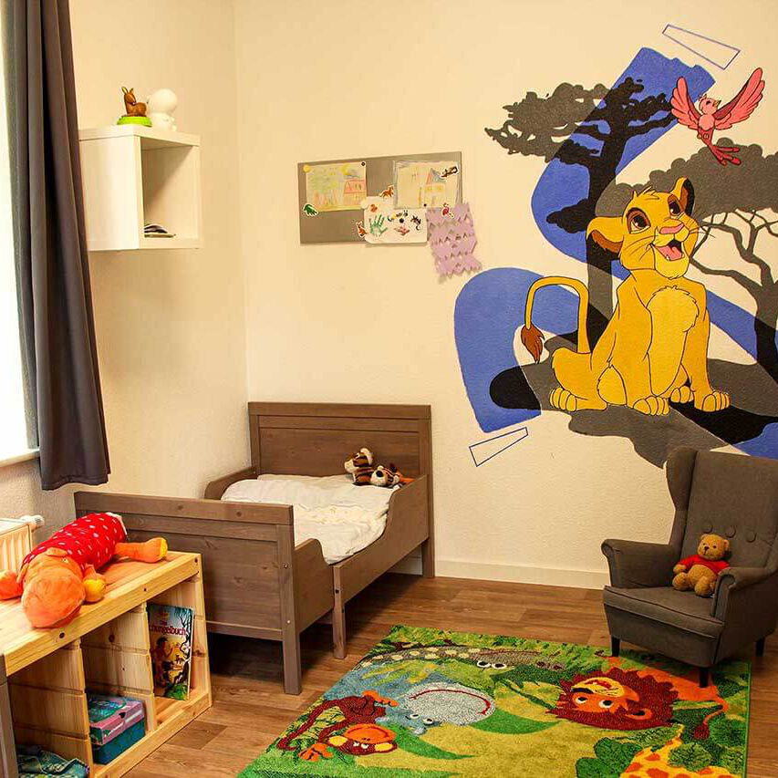 Ein Doppelzimmer mit zwei Kinderbetten, einem Regal, Spielteppich, Sessel und einer Wandbemalung König der Löwen.