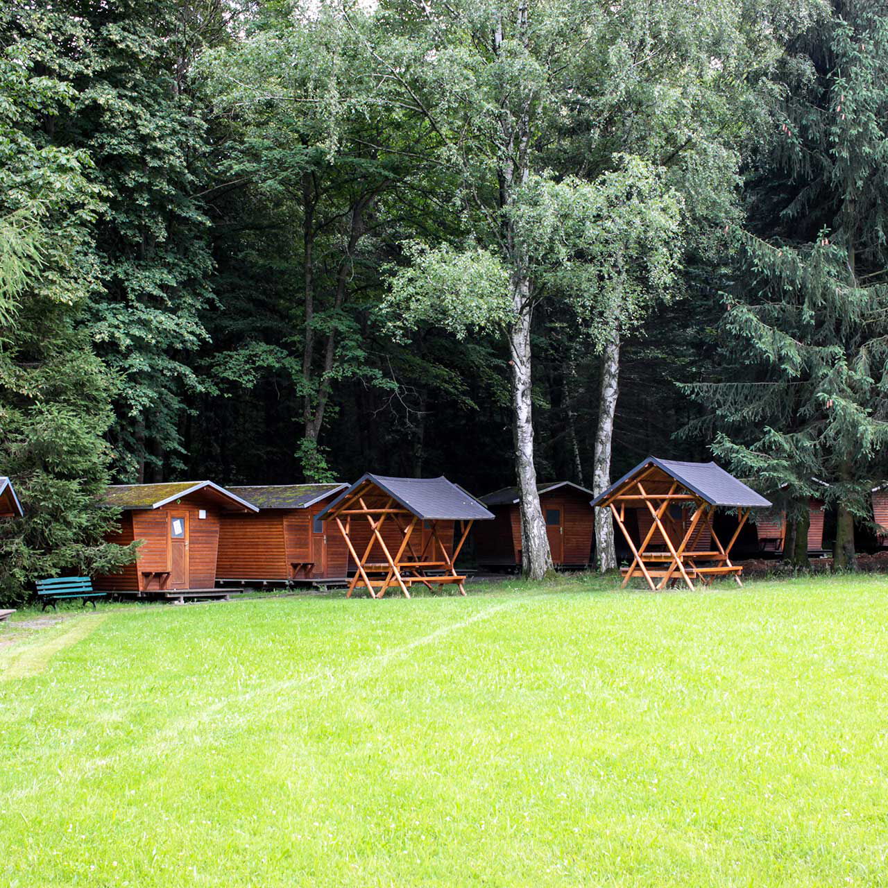 Zehn Abenteuer-Holzhäuschen am Waldrand, eine große Wiese und zwei überdachte Sitzgruppen.