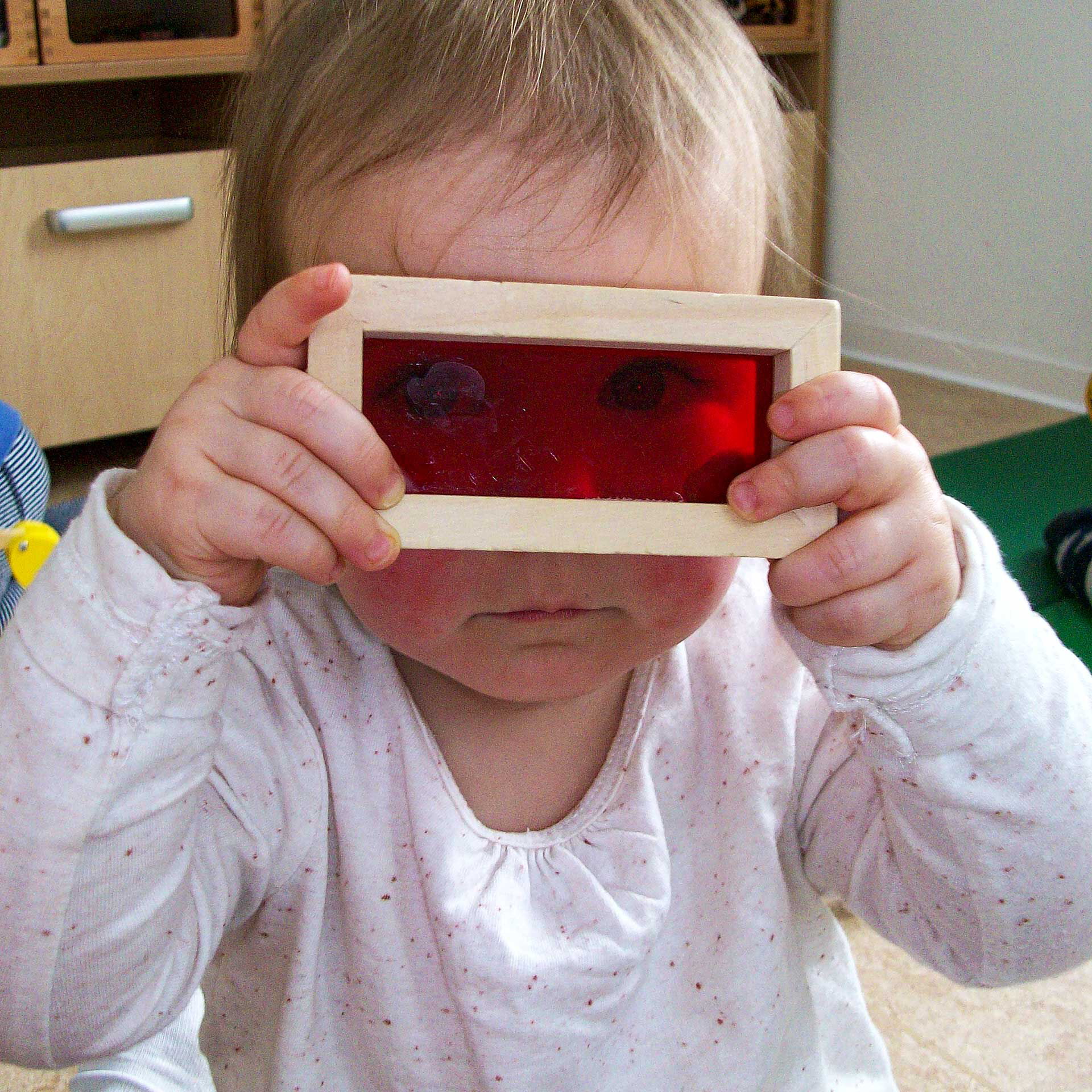 Ein Kleinkind hält sich ein Baustein vor die Augen. In der Mitte ist der Holzbaustein mit einer roten Folie durchsichtig.