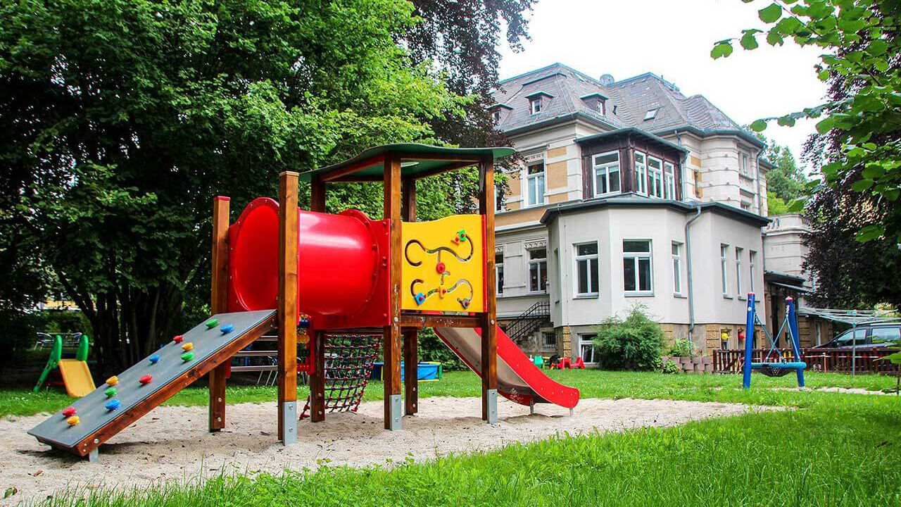 Der Spielplatz im Außengelände mit einem Klettergerüst mit Rutsche und einer Nestschaukel. Im Hintergrund ist die Villa.