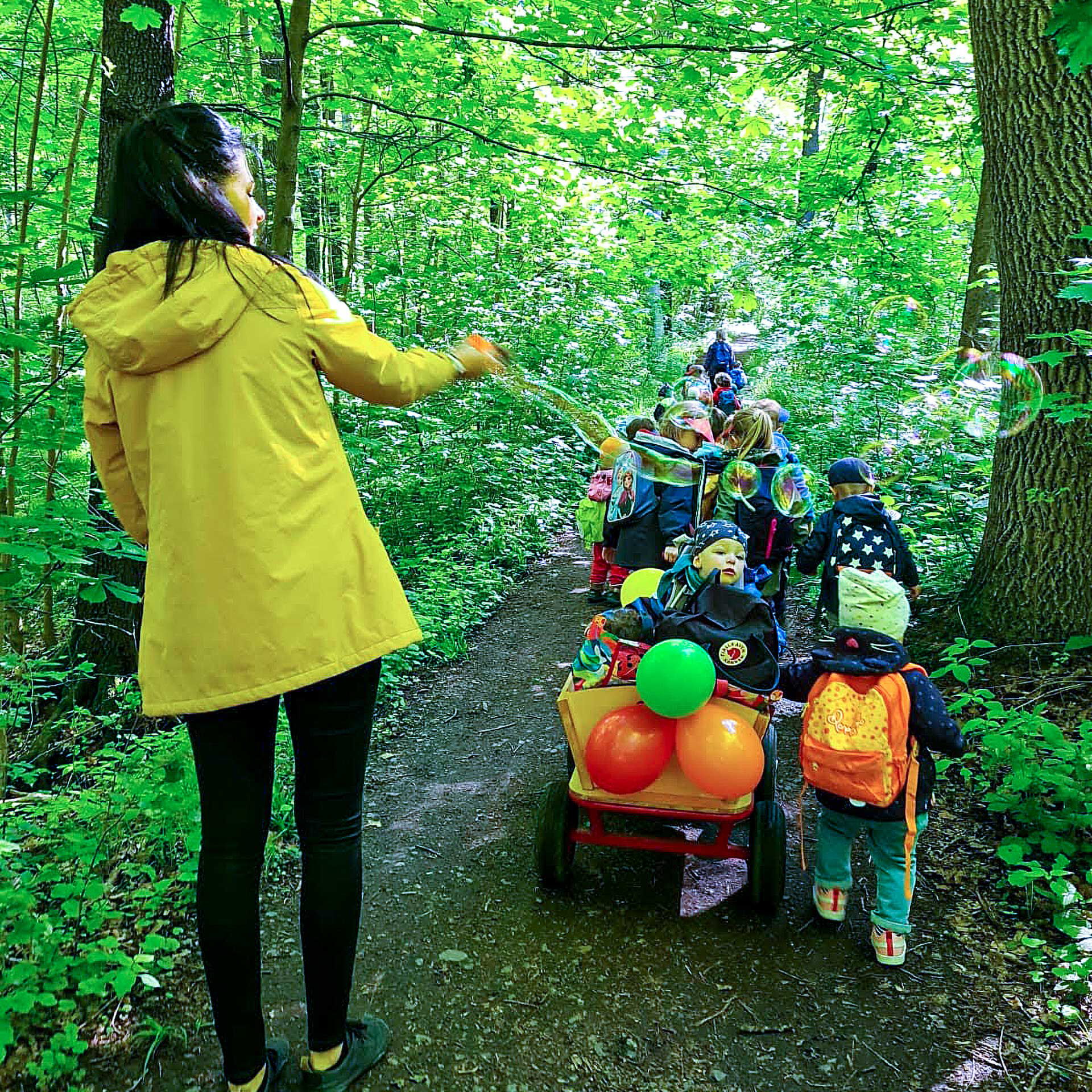 Ein Ausflug einer Kindergruppe in den Wald. Die Erzieherin lässt Seifenblasen fliegen und Kinder sitzen im Bollerwagen oder laufen nebenher.