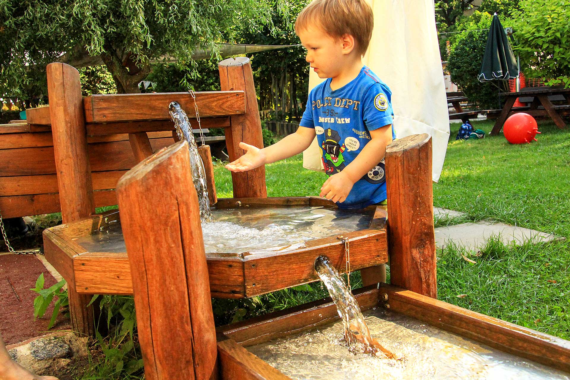 Die Matschanlage mit Wasserspiel aus Holz im Außengelände. Ein Junge spielt mit dem Wasser.